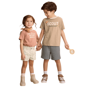 Two children hold hands, smiling. The girl wears a peach shirt with \“Let's Use Our Outside Voices,\“ white shorts, and beige boots. The boy wears a tan \“Scout\“ shirt, gray shorts, and brown sneakers, holding a wooden magnifying glass.