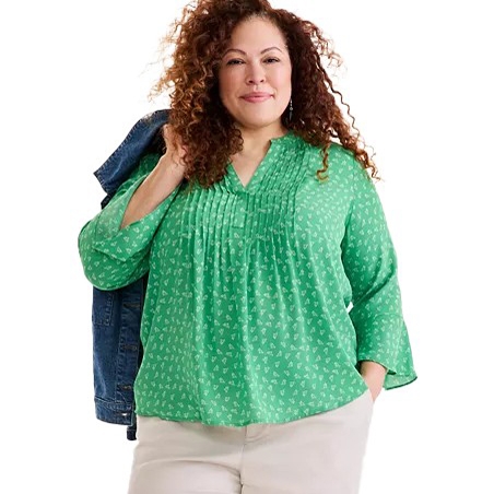 A woman wearing a light green blouse with a subtle pattern and a relaxed fit, paired with light-colored pants. She's carrying a denim jacket over her shoulder.