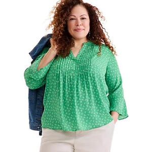 A woman wearing a light green blouse with a subtle pattern and a relaxed fit, paired with light-colored pants. She's carrying a denim jacket over her shoulder.
