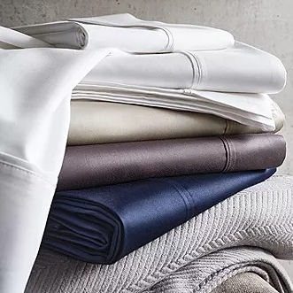 A stack of neatly folded sheets in various colors, including white, beige, brown, and navy, resting on a light-colored textured fabric.