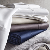 A stack of neatly folded sheets in various colors, including white, beige, brown, and navy, resting on a light-colored textured fabric.