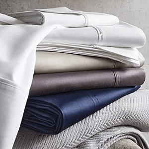 A stack of neatly folded sheets in various colors, including white, beige, brown, and navy, resting on a light-colored textured fabric.