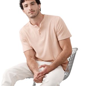A man is seated on a metal chair, wearing a light peach short-sleeve henley shirt and white pants.