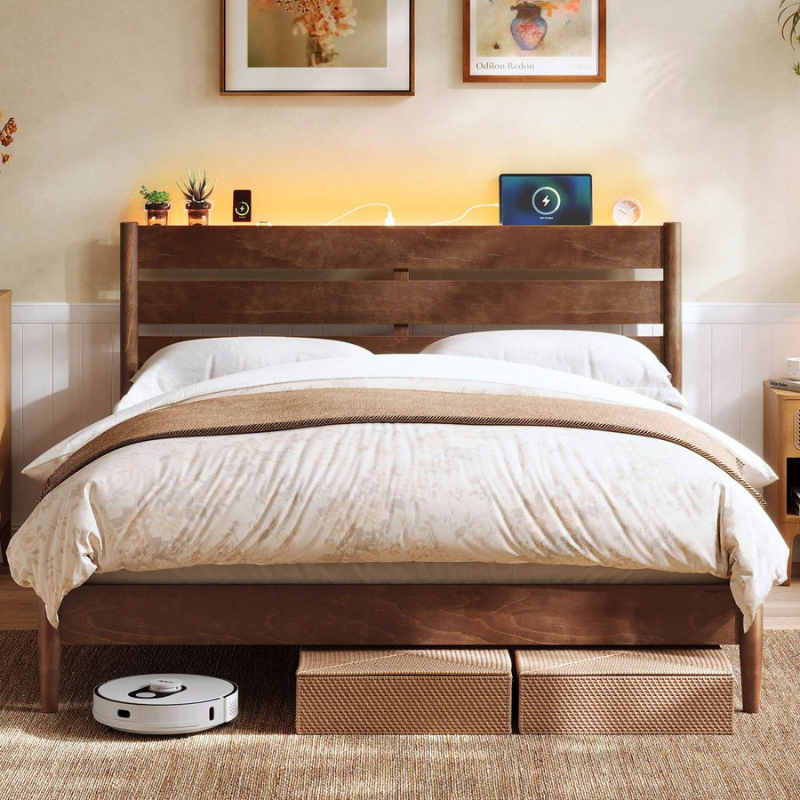 A bedroom scene featuring a wooden bed frame with a robotic vacuum cleaner underneath, two storage boxes, a pair of charging devices on the headboard, and framed artwork on the wall.