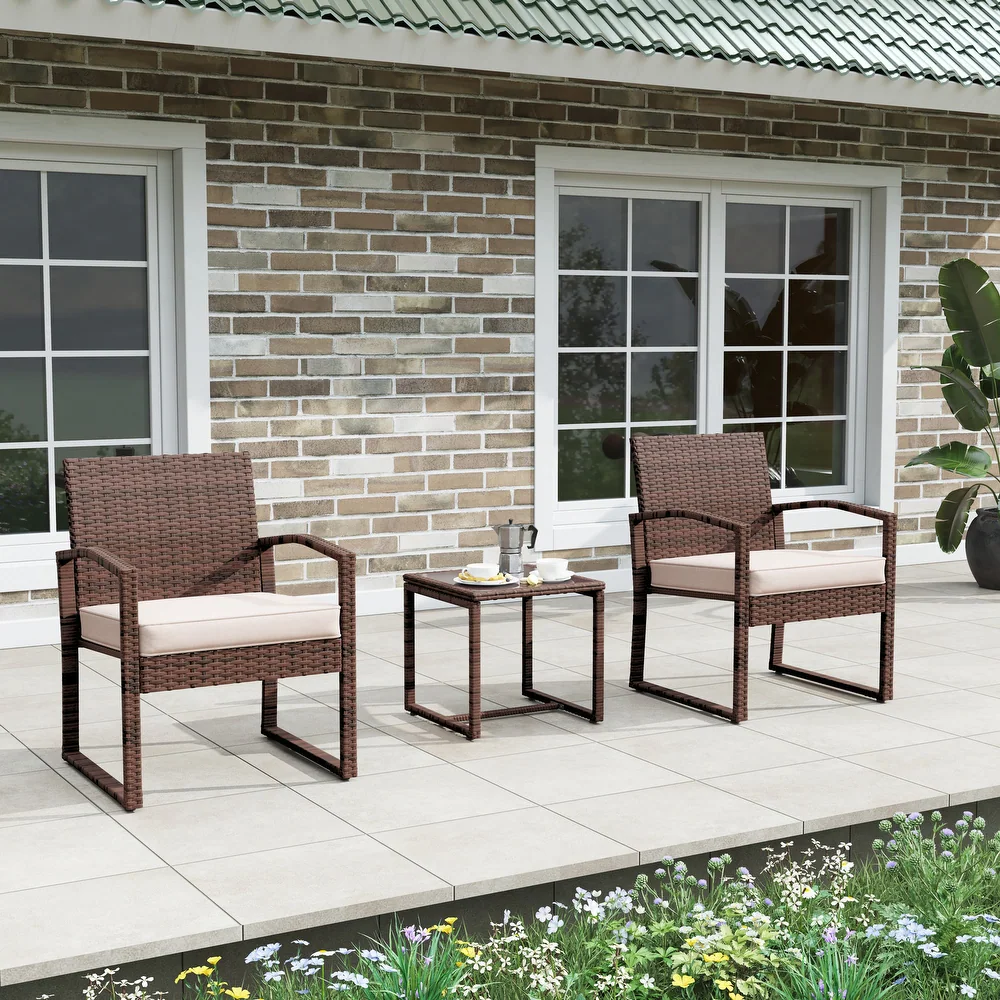Rattan-style chat set with two cushioned chairs and a square side table in brown wicker, set on a tiled patio with a brick wall backdrop.