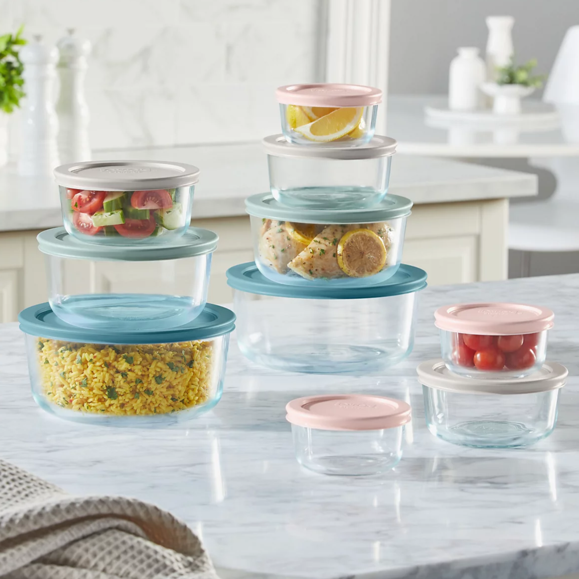 Stacked, round glass storage containers with pastel-colored lids, some holding food items like salad, lemon wedges, rice, and tomatoes, are arranged on a kitchen countertop.