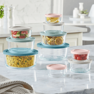 Stacked, round glass storage containers with pastel-colored lids, some holding food items like salad, lemon wedges, rice, and tomatoes, are arranged on a kitchen countertop.