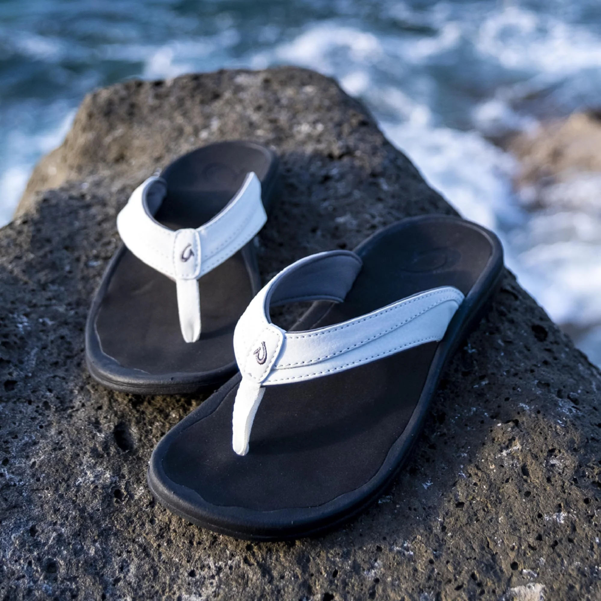 White and black flip-flops with a sleek design placed on a rocky surface against a background of ocean waves.