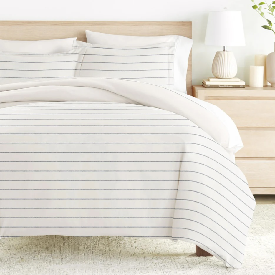 A neatly arranged bed with a striped white duvet cover and matching pillows, accompanied by a light wood nightstand featuring a white lamp and decorative vases.