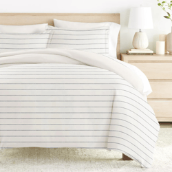 A neatly arranged bed with a striped white duvet cover and matching pillows, accompanied by a light wood nightstand featuring a white lamp and decorative vases.