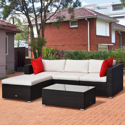 Outdoor patio furniture set with a black wicker frame, beige cushions, and red accent pillows, featuring a sectional sofa and a rectangular coffee table on a brick patio.