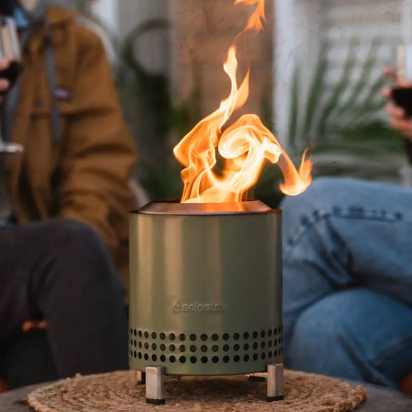 A portable camping stove with a visible flame is on display, set on a woven mat.