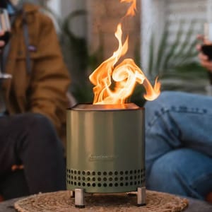 A portable camping stove with a visible flame is on display, set on a woven mat.