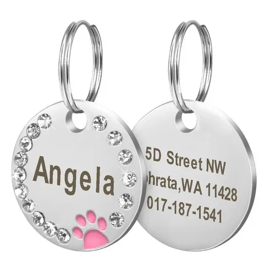 Two round pet ID tags with silver tones and rhinestone embellishments, showcasing the name \“Angela\“ and a pink paw print on the front. The back displays an address and contact number.