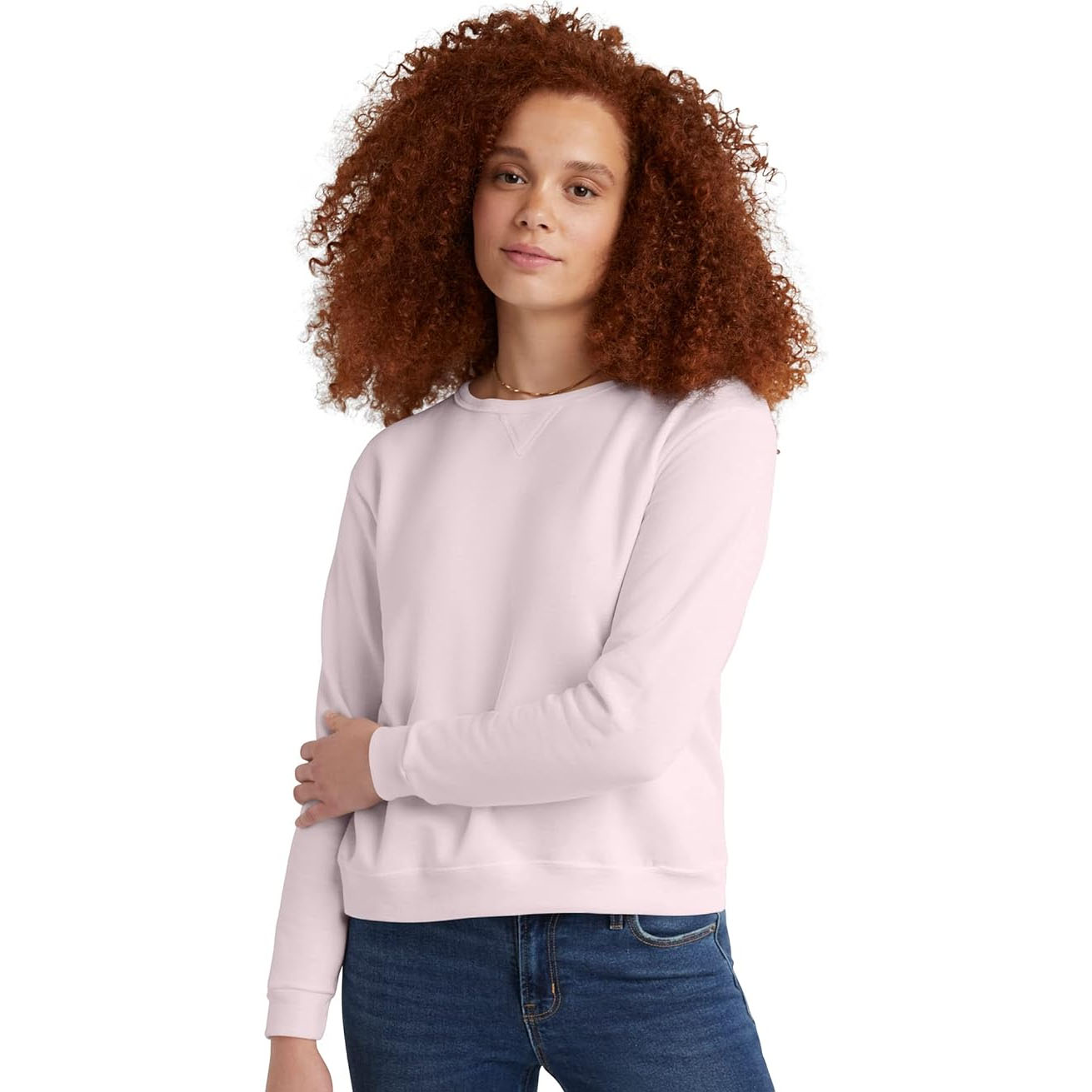 A woman with curly hair is wearing a light pink Hanes sweatshirt featuring a simple crew neck and long sleeves, paired with blue jeans.