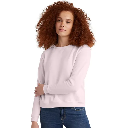 A woman with curly hair is wearing a light pink Hanes sweatshirt featuring a simple crew neck and long sleeves, paired with blue jeans.