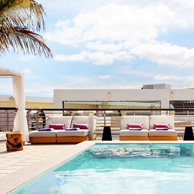 Outdoor pool area with cushioned lounge chairs featuring pink and purple pillows under a partly cloudy sky. A palm tree and a white structure with horizontal railings are in the background.