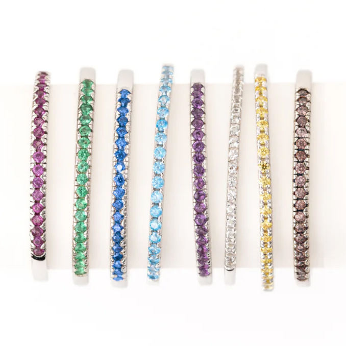 A collection of eight thin bangles, each set with round gemstones in different colors: purple, green, blue, aquamarine, dark purple, white, yellow, and brown, all in a white metal setting.