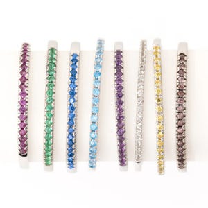 A collection of eight thin bangles, each set with round gemstones in different colors: purple, green, blue, aquamarine, dark purple, white, yellow, and brown, all in a white metal setting.