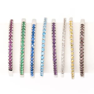 A collection of eight thin bangles, each set with round gemstones in different colors: purple, green, blue, aquamarine, dark purple, white, yellow, and brown, all in a white metal setting.