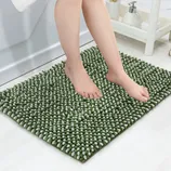 A pair of feet standing on a green, textured bath mat with a chenille noodle design, placed on a white bathroom floor. Soap dispenser and towel rack are visible in the background.