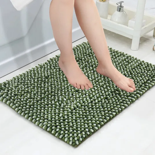 A pair of feet standing on a green, textured bath mat with a chenille noodle design, placed on a white bathroom floor. Soap dispenser and towel rack are visible in the background.