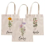 Three beige tote bags with floral designs and personalized names: Amanda, Emily, and Angela. Each bag features a different flower illustration in the center.