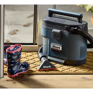 A Shark MessMaster vacuum cleaner is pictured cleaning a doormat; its compact design, blue and gray color scheme, built-in handle, and flexible hose are visible. Nearby is a pair of children's rain boots.