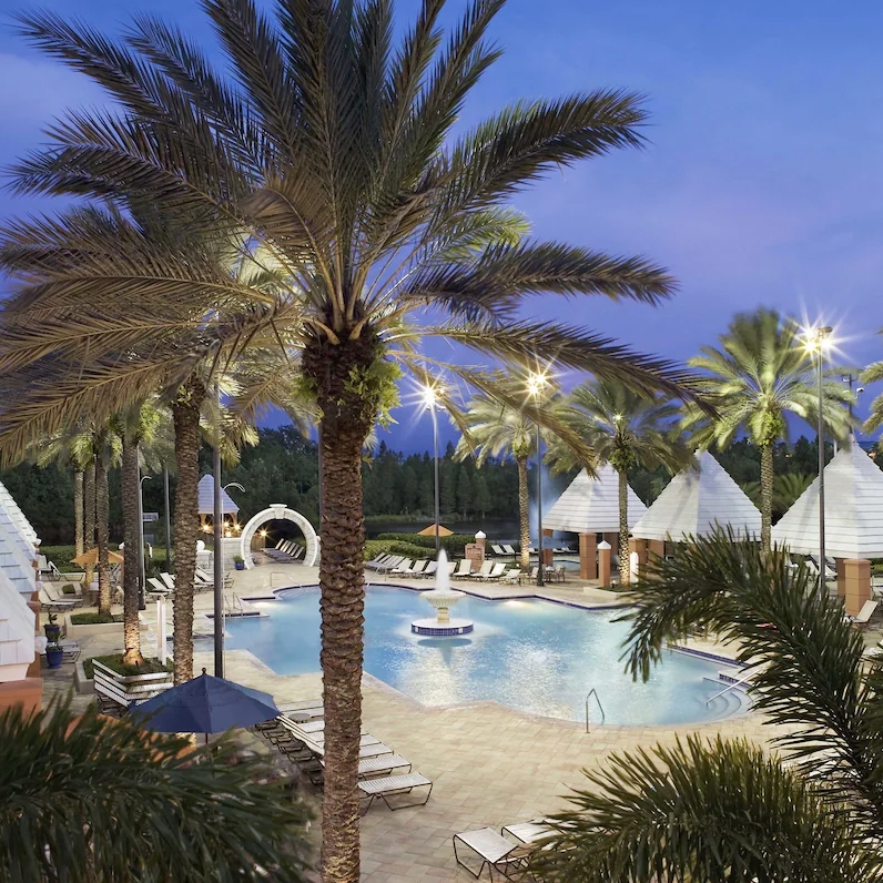 A luxurious pool area surrounded by palm trees and modern, geometric cabanas, with lounge chairs and a tranquil ambiance under evening lights.