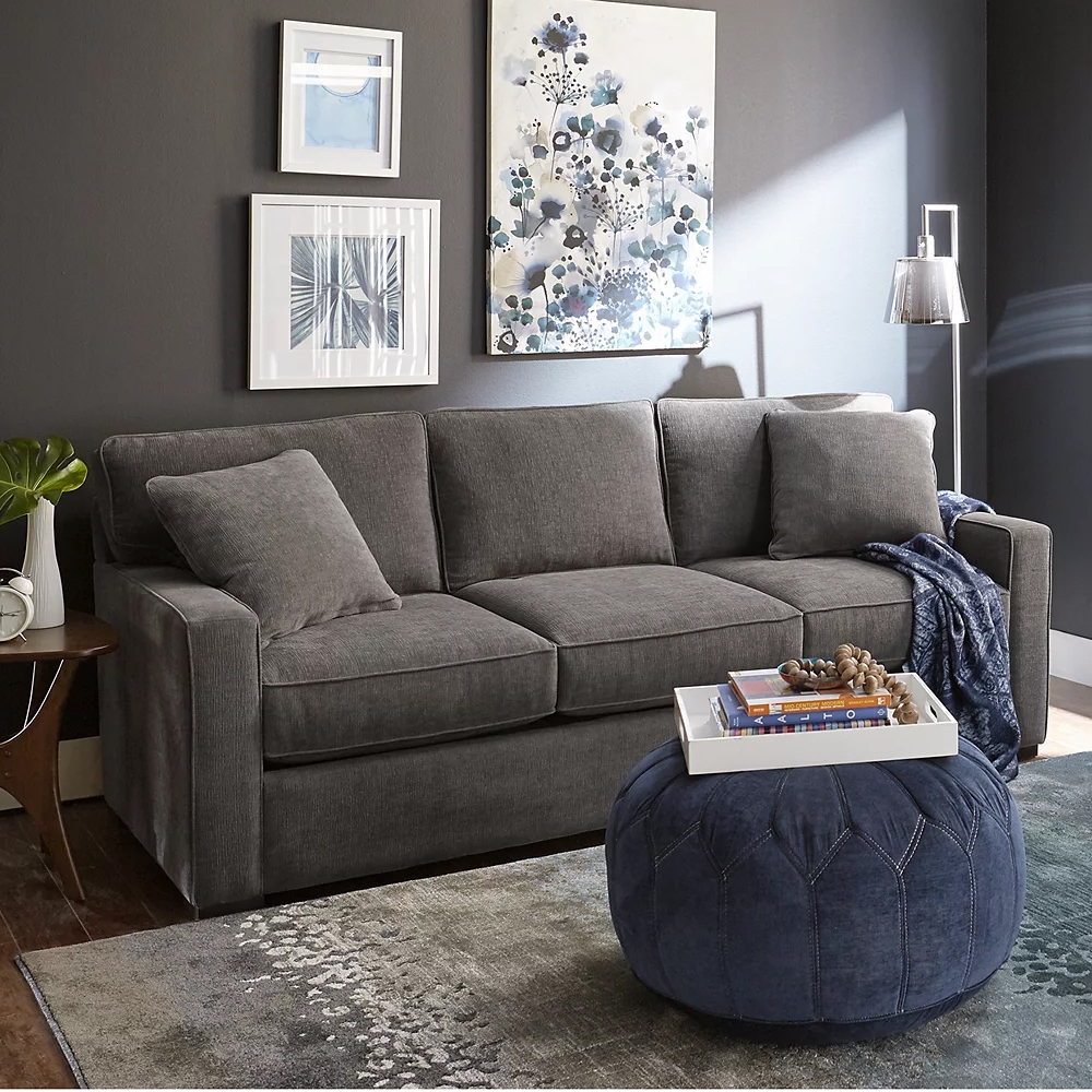 A 62-inch grey fabric loveseat with simple cushions and straight arms, set against a dark wall with framed art. A pouf with a tray sits in front, and a floor lamp provides light.