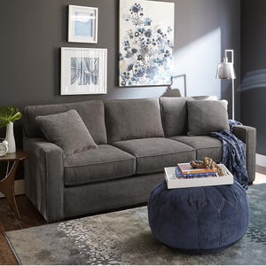 A 62-inch grey fabric loveseat with simple cushions and straight arms, set against a dark wall with framed art. A pouf with a tray sits in front, and a floor lamp provides light.