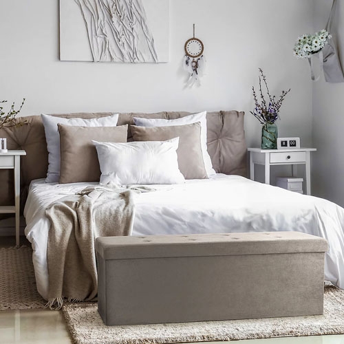 A neatly made bed with beige and white bedding and a gray bench at the foot, set in a bedroom with simple decor.