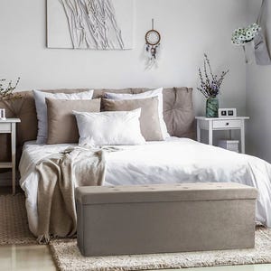 A neatly made bed with beige and white bedding and a gray bench at the foot, set in a bedroom with simple decor.