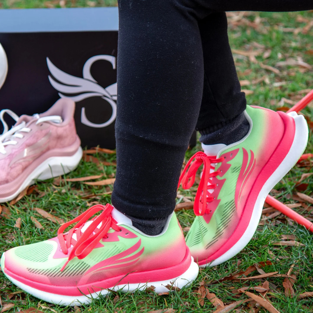 Brightly colored running shoes with red laces are worn on grass, next to a box and a pair of pink sneakers.