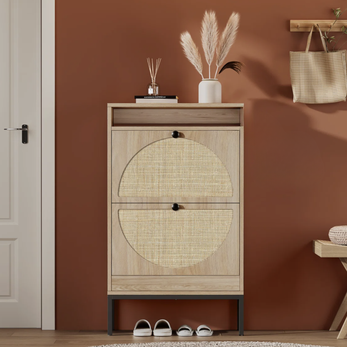 A wooden shoe cabinet with two woven-patterned doors stands against a terracotta wall. It features decor items like a vase with pampas grass on top, shoes underneath, and nearby a bench and a hanging bag.
