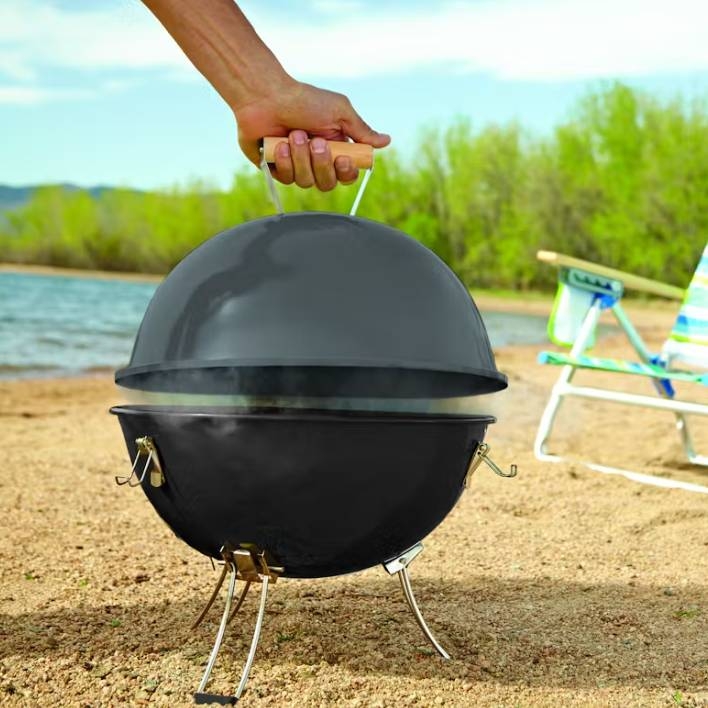 A portable charcoal grill with a round, black lid, held by a person's hand, set outdoors near a body of water and lounge chairs.