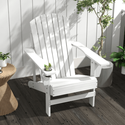 White Adirondack chair with built-in cup holder, placed on a wooden deck next to a rattan side table topped with a plant and book; surrounded by additional potted greenery.