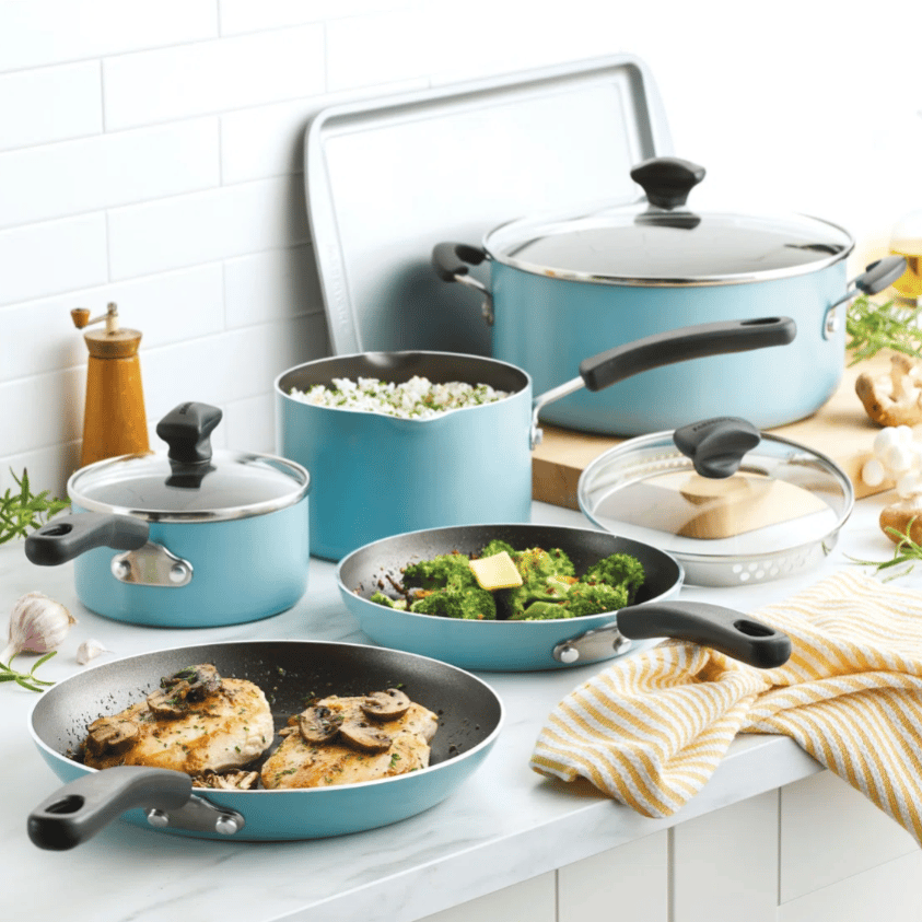 A set of light blue non-stick cookware with black handles, including frying pans with cooked chicken and broccoli, pots with lids, a large pot, and a baking sheet, arranged on a white kitchen counter.