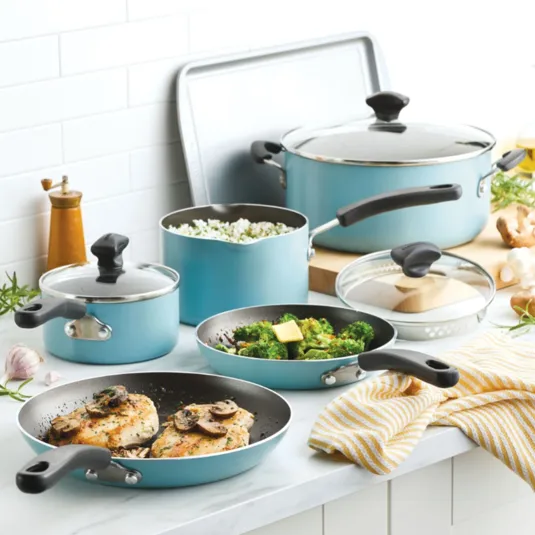 A set of light blue non-stick cookware with black handles, including frying pans with cooked chicken and broccoli, pots with lids, a large pot, and a baking sheet, arranged on a white kitchen counter.
