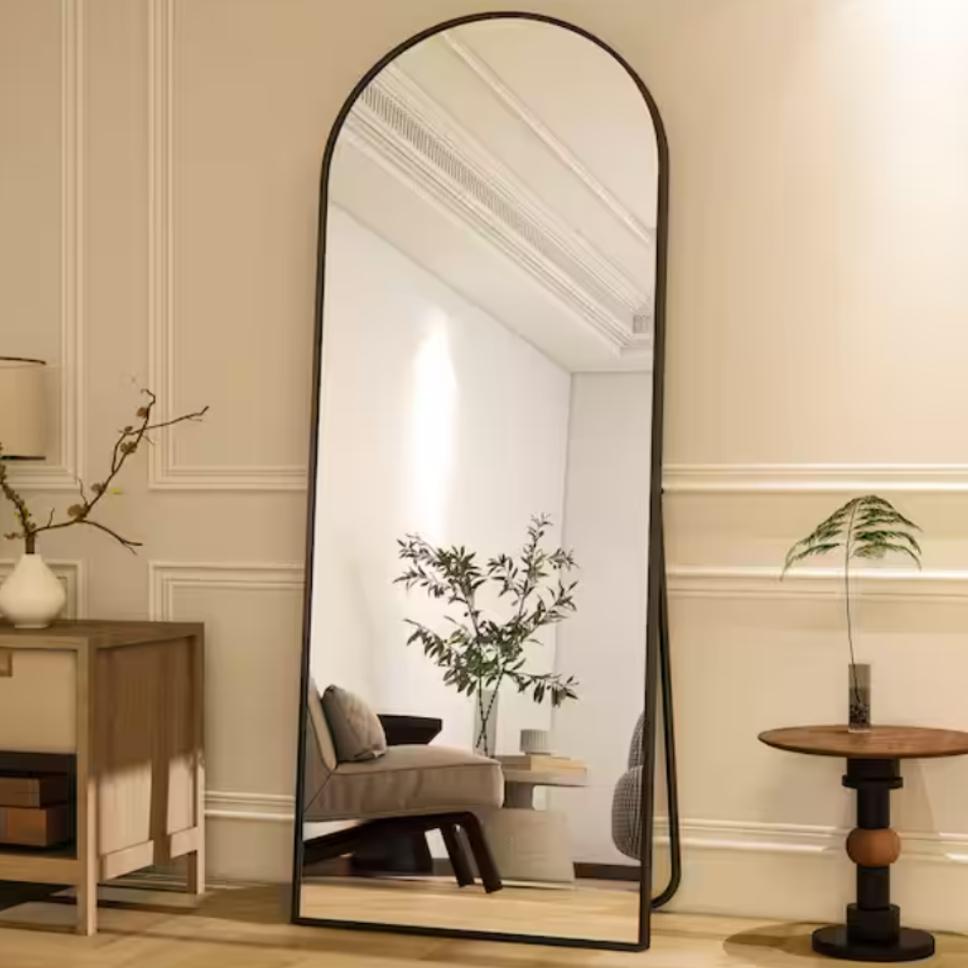 A tall, arched floor mirror with a black frame is leaning against a wall in a room featuring a side table, potted plants, and a decorative lamp.