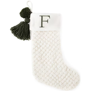 A white knitted Christmas stocking with a textured pattern features a green \“F\“ monogram on the cuff and is decorated with a green pom-pom and tassel.
