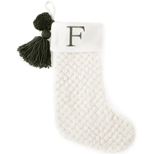 A white knitted Christmas stocking with a textured pattern features a green \“F\“ monogram on the cuff and is decorated with a green pom-pom and tassel.
