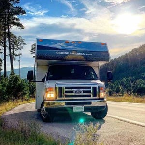 An RV from Cruise America is parked on a scenic road surrounded by trees and hills, with a clear blue sky above.