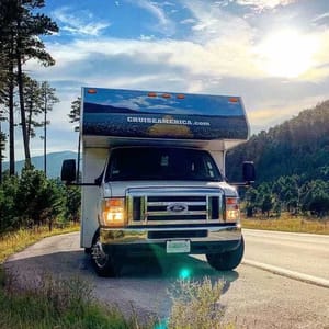 An RV from Cruise America is parked on a scenic road surrounded by trees and hills, with a clear blue sky above.