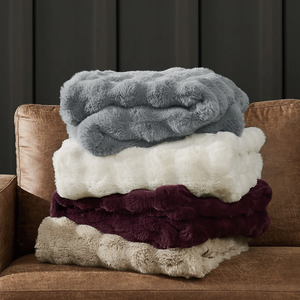 A stack of four plush, fluffy blankets in gray, white, purple, and beige sits on a brown leather chair.
