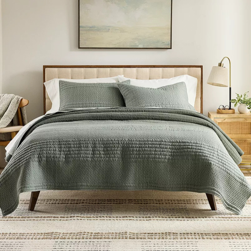 A neatly made bed with a green quilt and matching pillows, accompanied by a wooden nightstand with a lamp and plant. A simple framed painting hangs above the bed.
