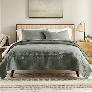 A neatly made bed with a green quilt and matching pillows, accompanied by a wooden nightstand with a lamp and plant. A simple framed painting hangs above the bed.
