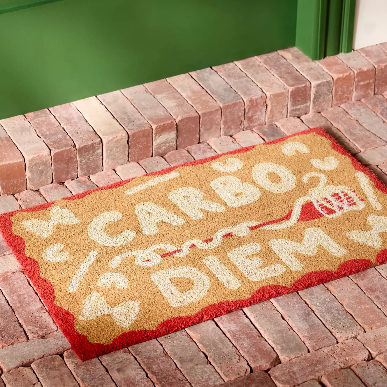 Doormat with the phrase \“CARBO DIEM\“ in playful lettering, featuring pasta illustrations, placed on a brick porch in front of a green door.