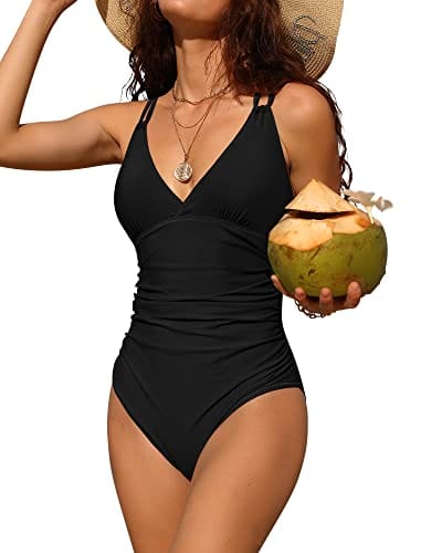 A woman is wearing a black one-piece swimsuit with a V-neckline and ruched waist for tummy control, holding a coconut.