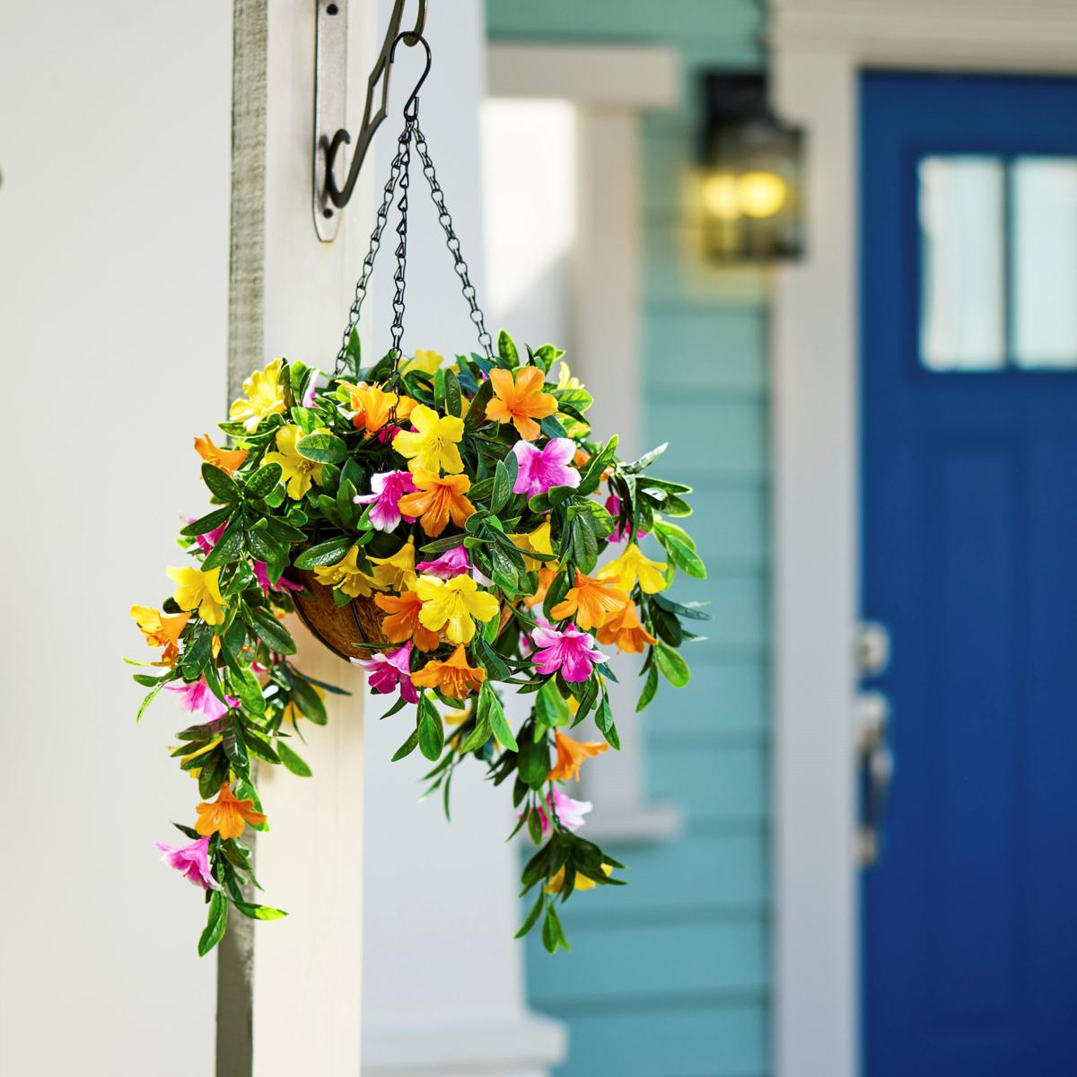 A set of two faux-azalea hanging baskets featuring vibrant yellow, orange, and pink flowers with green foliage, suspended by a chain against a backdrop of a light blue house and a navy blue door.